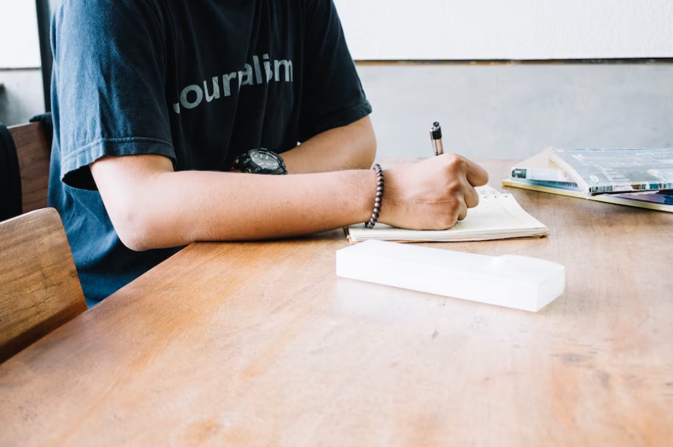 The person in a black t-shirt  sits at the table and makes notes with a pen