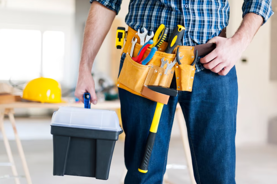 man in jeans holding equipment for repair, tools in his pocket, the yellow helmet behind